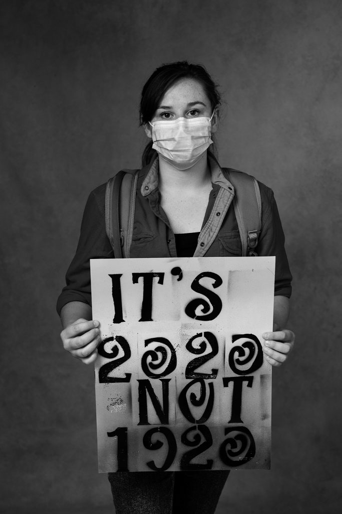 Black and white portrait of a woman wearing a face mask and holding a sign that reads "It's 2020, not 1920"