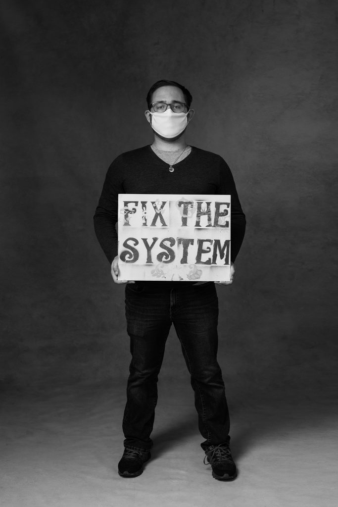 Black and white portrait of man wearing mask and holding a sign that reads "Fix the System"
