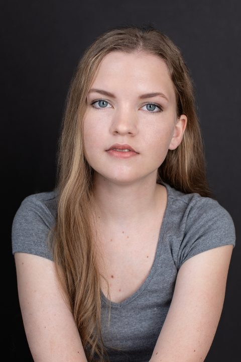 Vertical headshot of Chloe, a young actor, wearing a gray t-shirt and photographed on a black background