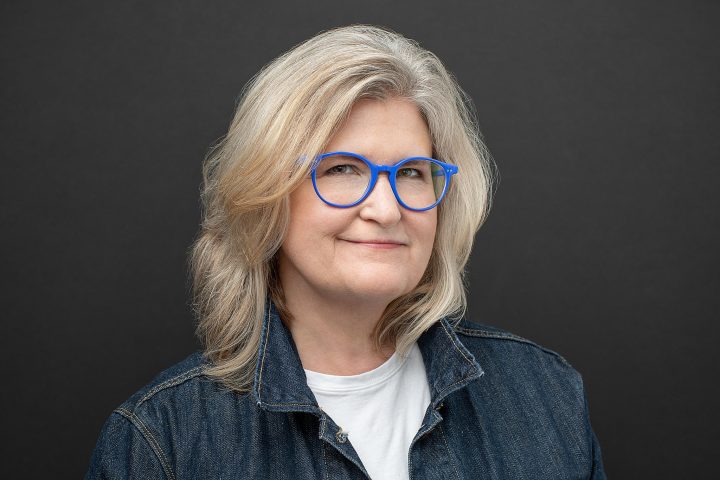 Headshot of woman with blue glasses and a denim jacket on a black background