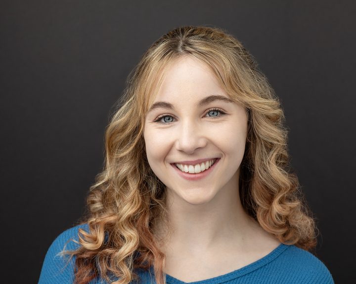 Acting headshot with a black background - Lily, smiling, wearing a blue sweater