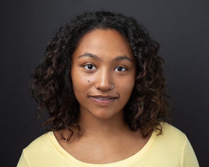 Headshot of singer / actor Janaysia Gethers, wearing yellow, with a dark background