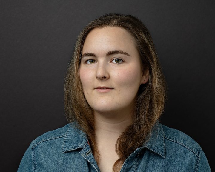 Acting headshot for Anna, who is wearing a jean jacket with her hair tucked in, on a black background