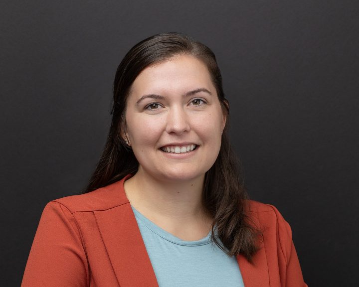 A business casual headshot of a woman in front of a black background, wearing an orange jacket
