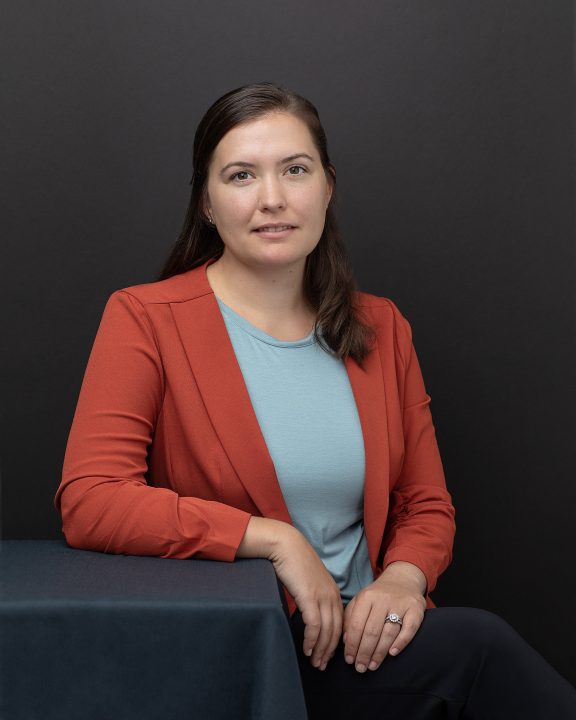 A business casual portrait of a woman sitting beside a table in front of a black background, wearing an orange jacket
