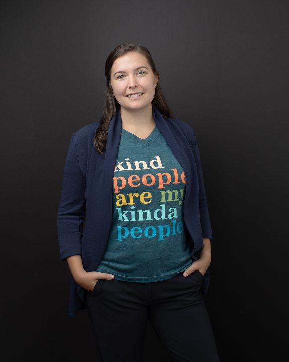 A casual portrait of a woman standing in front of a black background, wearing a t-shirt