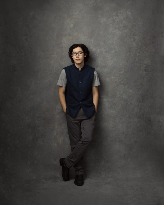 A casual portrait of a 16-year-old boy, leaning against a gray, hand-painted backdrop, wearing a blue waistcoat