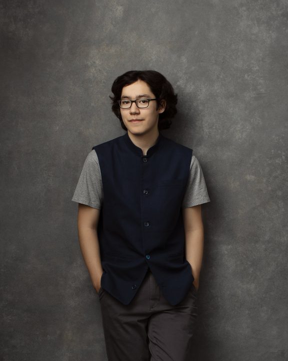 A casual portrait of a 16-year-old boy, leaning against a gray, hand-painted backdrop, wearing a blue waistcoat