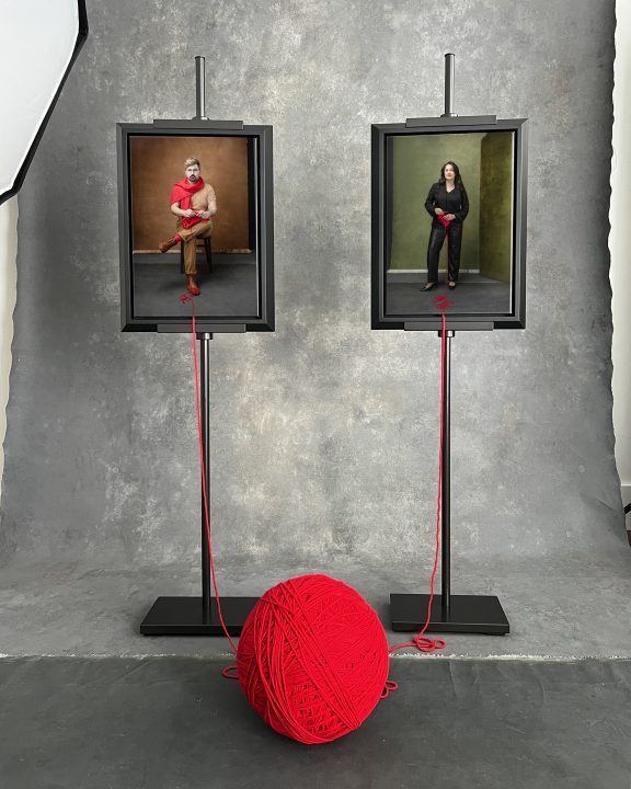A cell phone photo of the Knitted Together project beginning, with two portraits printed on canvas, framed, on easels, with strands of yarn connecting them to a large ball.