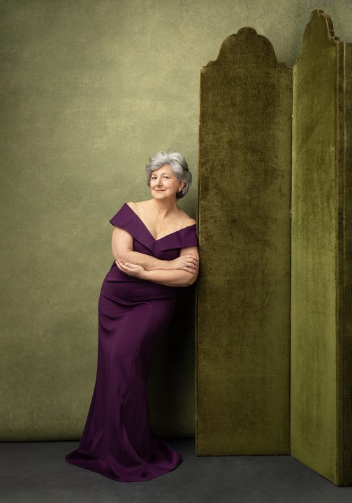 Portrait of a woman in her 70s, wearing a purple dress in front of a green background