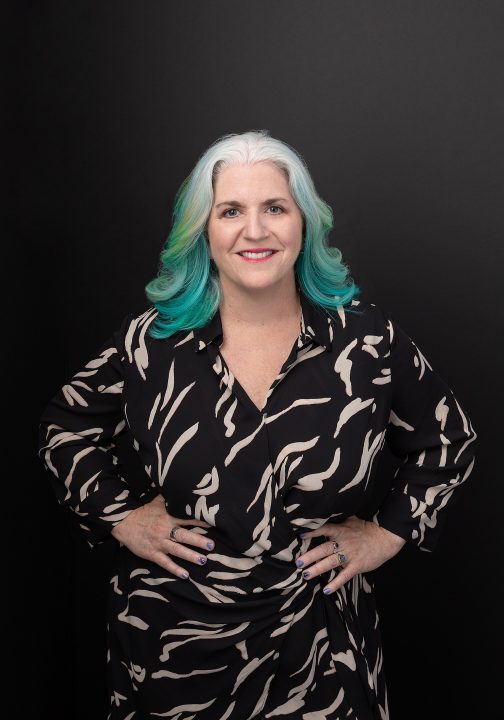 Portrait of Kim smiling, wearing a cream/black patterned dress, in front of black background