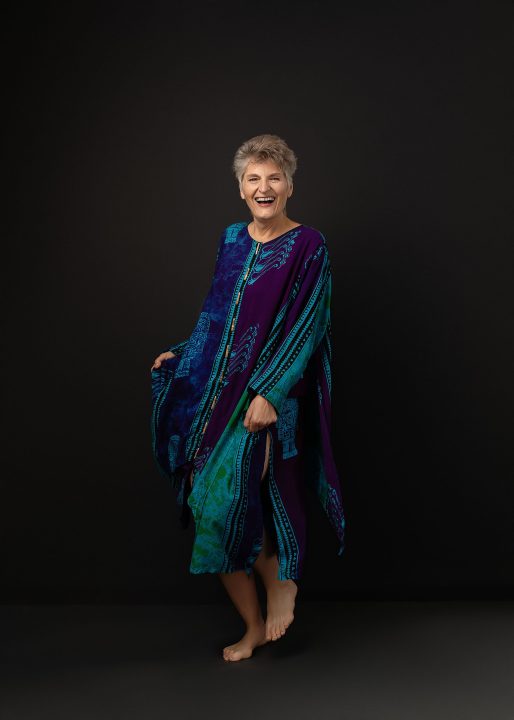 A portrait of Sue from her photo session for The Over 50 Revolution. She is wearing a colorful dress, dancing in front of a black background.