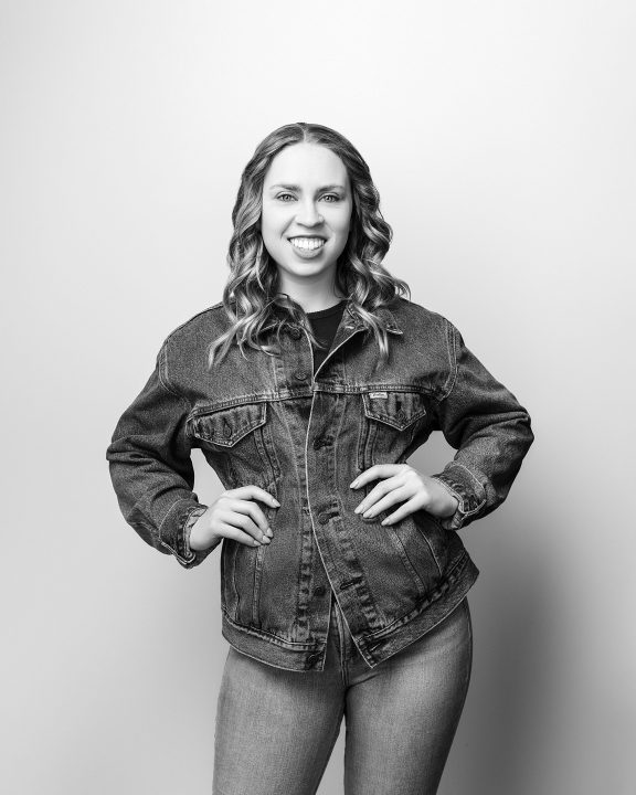 A black and white portrait of Kenzie from her five year update, smiling, in front of a light backdrop. She is wearing a denim jacket and jeans.
