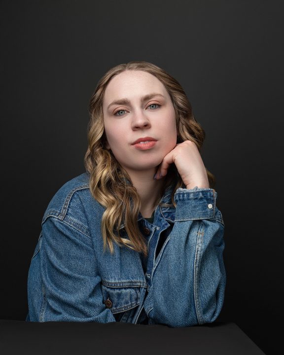 A photo from Kenzie's five year update. She is seated and wearing a denim jacket in front of a black background.