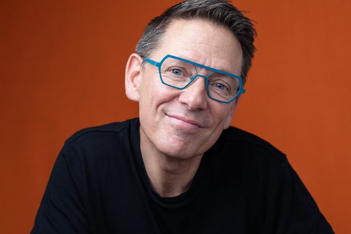 Portrait of Dan Perkins for Artisan Eyewear.  Wearing bold teal frames.  Bright orange background.