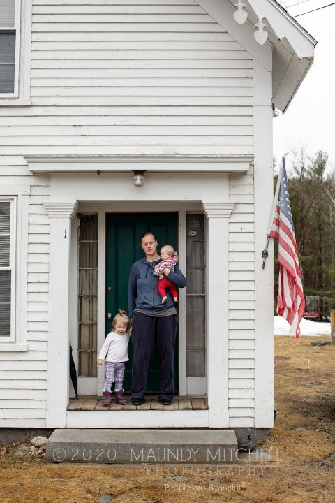 Amy, Molly, and baby Iris in the doorway of their home, next to a worn American flag