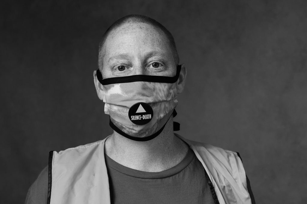 Closeup portrait of woman with shaved head, wearing "Silence=Death" badge on her mask