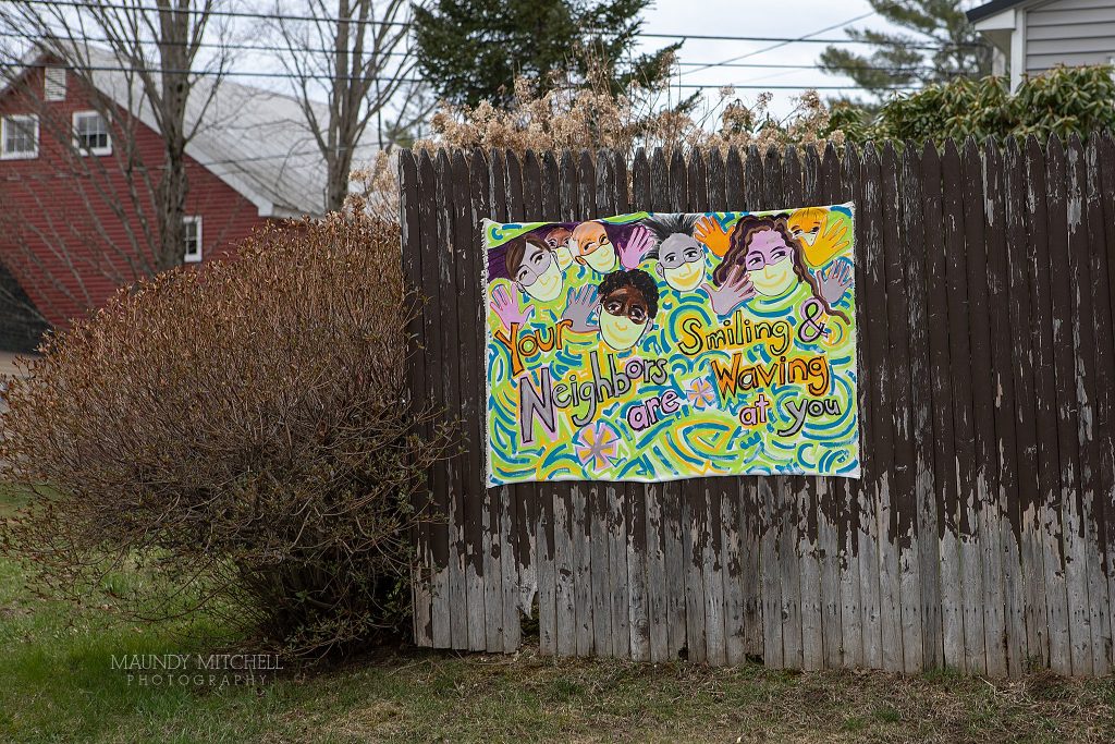 A hand painted sign on a fence reads "your neighbors are smiling and waving at you" by artist Marcia Santore