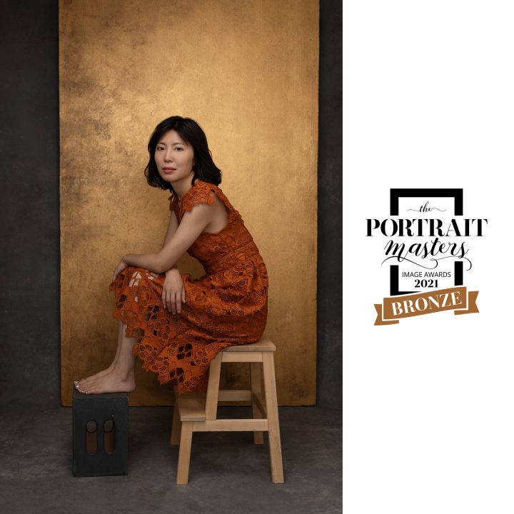 Portrait of Zi, seated, wearing orange lace dress in front of metallic gold backdrop - Bronze Award