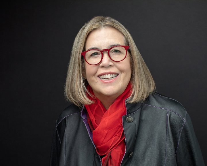 Headshot with dark background for NH artist and author Suzan Gannett, who is smiling and wearing red eyeglasses and a red scarf