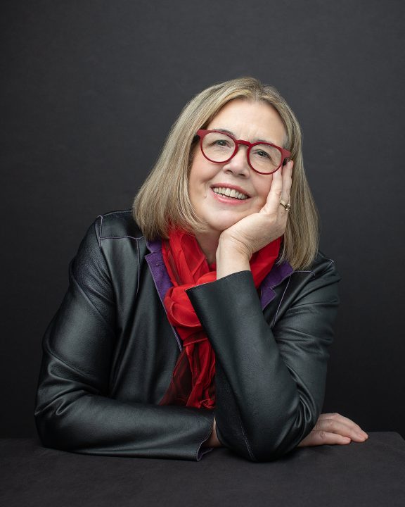 Headshot with dark background for NH artist and author Suzan Gannett, who is wearing red eyeglasses and a red scarf