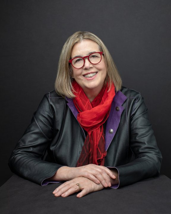 Headshot with dark background for NH artist and author Suzan Gannett, who is sitting at a table, wearing red eyeglasses and a red scarf