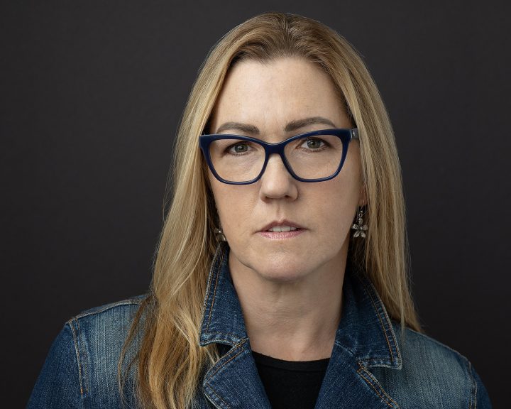 headshot of artist Amy Ford wearing a denim jacket and glasses with a black background
