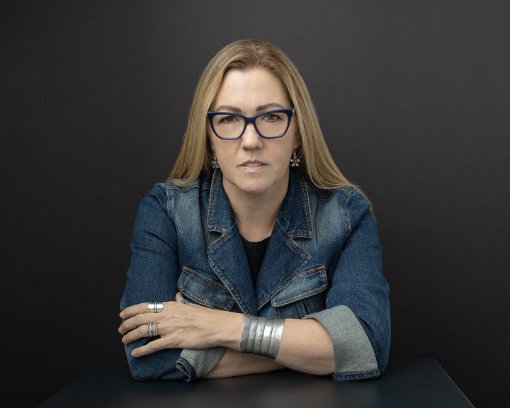 Artist Amy Ford, seated, wearing denim jacket - black background