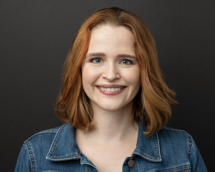 Professional headshot of Rebecca smiling, wearing a casual outfit with dark background
