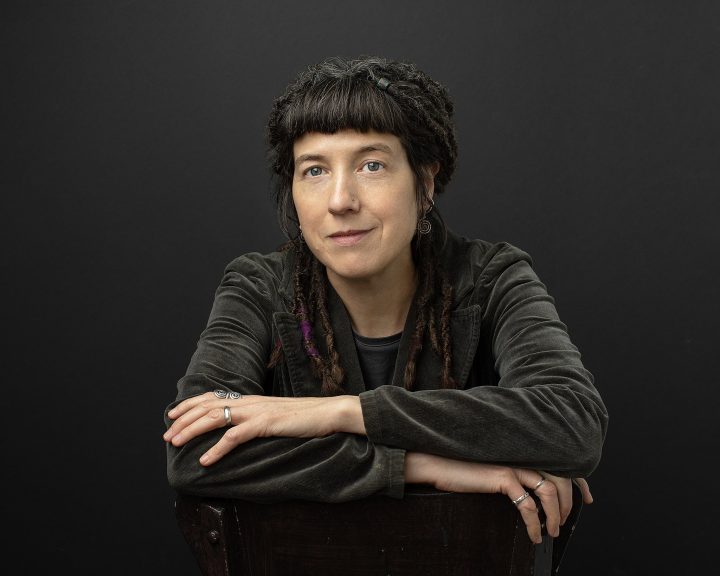 Portrait of Dr. Niki Tulk with dreadlocks, seated, arms crossed on the back of a chair, with a black background
