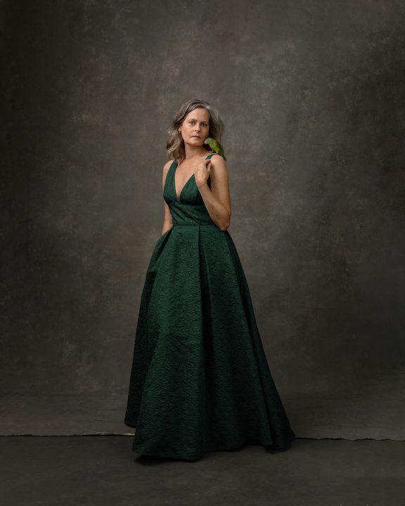 A full-length self-portrait of photographer Maundy Mitchell, wearing a green gown and holding a small parrot on her shoulder