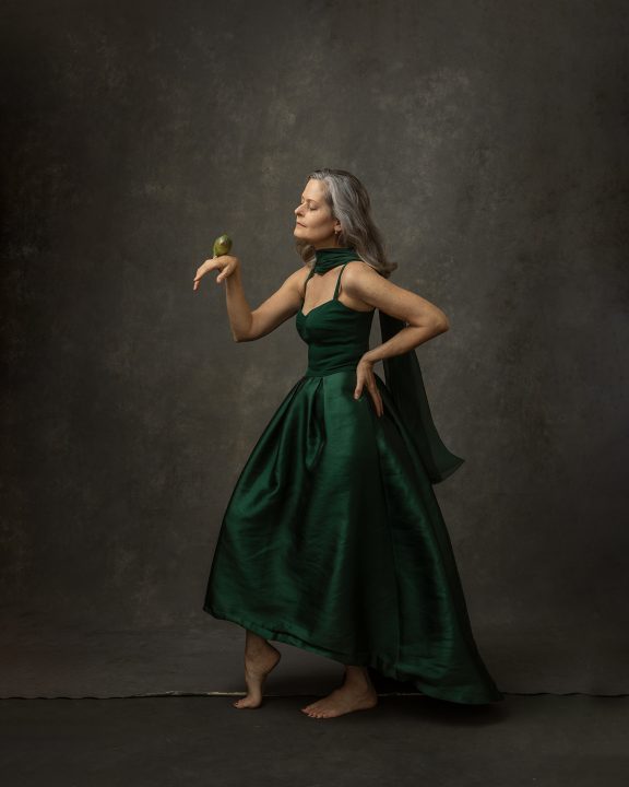 Self-portrait by Maundy Mitchell.  She is wearing a green gown and scarf, posing like a bird, and holding a little green parrot.