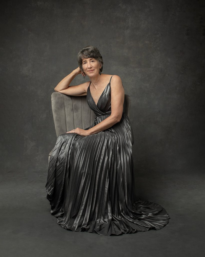 A monochromatic portrait of a woman in her 60s, relaxing in a chair, wearing a silver gown