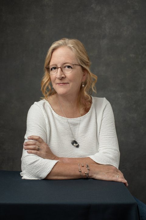 A casual portrait of a woman in her 60s, wearing a white shirt and glasses