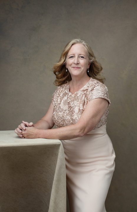 Portrait of a woman in her 60s, wearing a pink beaded gown, leaning on a table