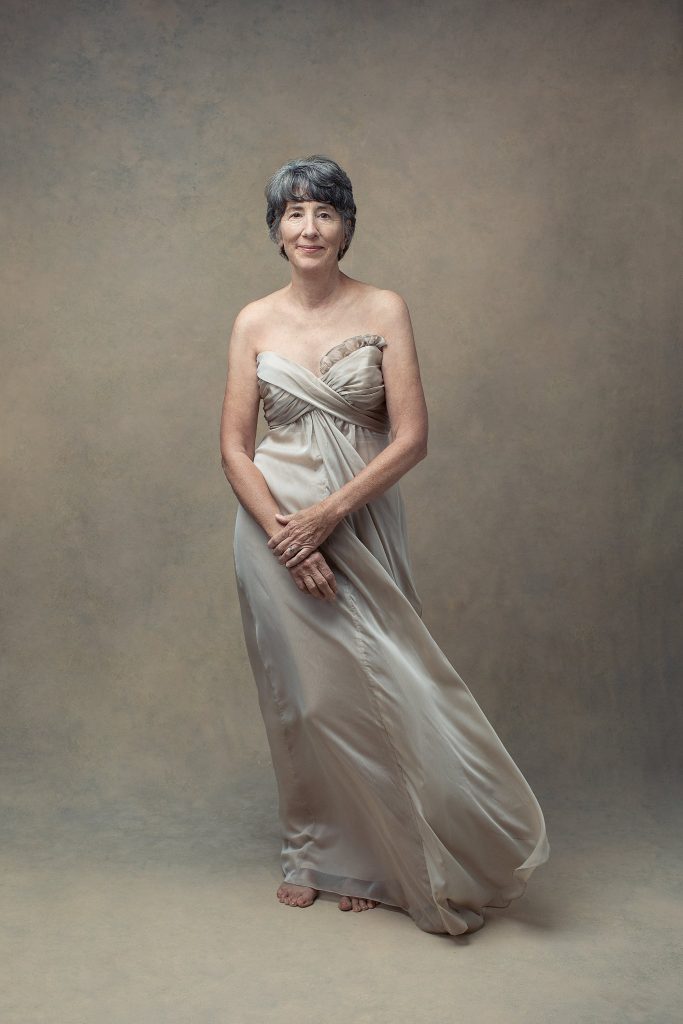 A monochromatic studio portrait of a woman in her 60s, wearing a light, flowy gown