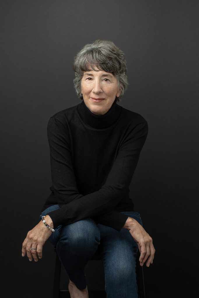 A casual studio portrait of a woman in her 60s, wearing a black turtleneck