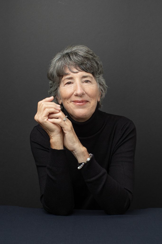 A portrait of a woman in her 60s, hands in frame, wearing a black turtleneck