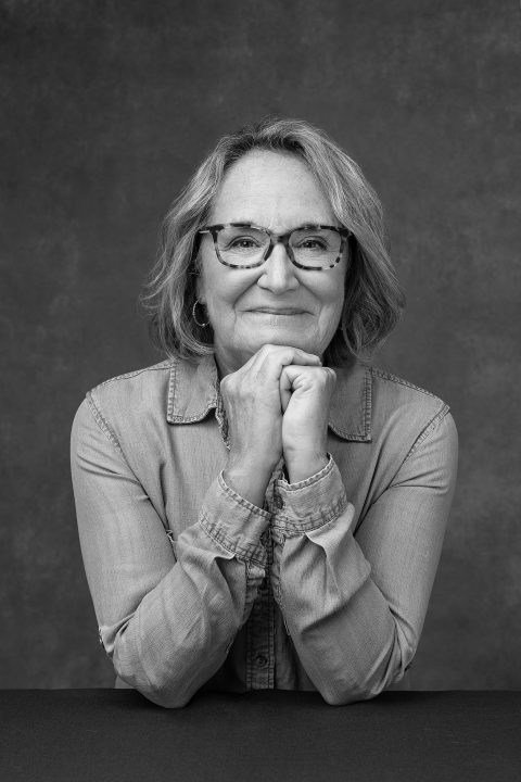 A black and white portrait of a woman in her 60s, wearing a denim shirt and glasses