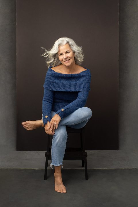 A casual portrait of a woman with white hair seated, barefoot, wearing a blue sweater and jeans in front of a layered backdrop