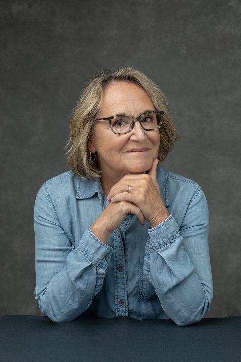 A casual portrait of a woman in her 60s, wearing a denim shirt and glasses