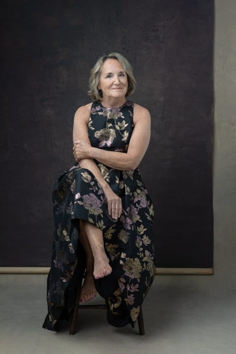 A portrait of a woman in her 60s, seated, wearing a floral metallic dress
