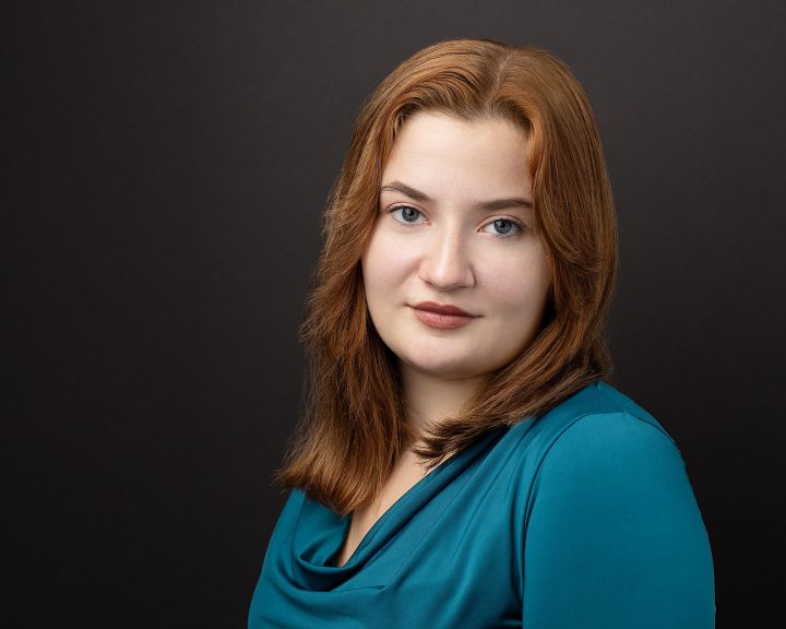 A headshot of an actor with red hair, a teal shirt, and a black background