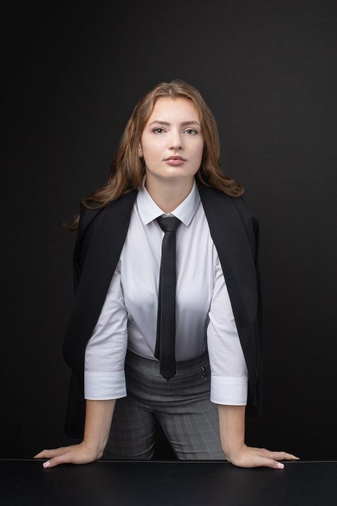 A high school senior girl wearing a shirt and tie, confidently leaning on a desk