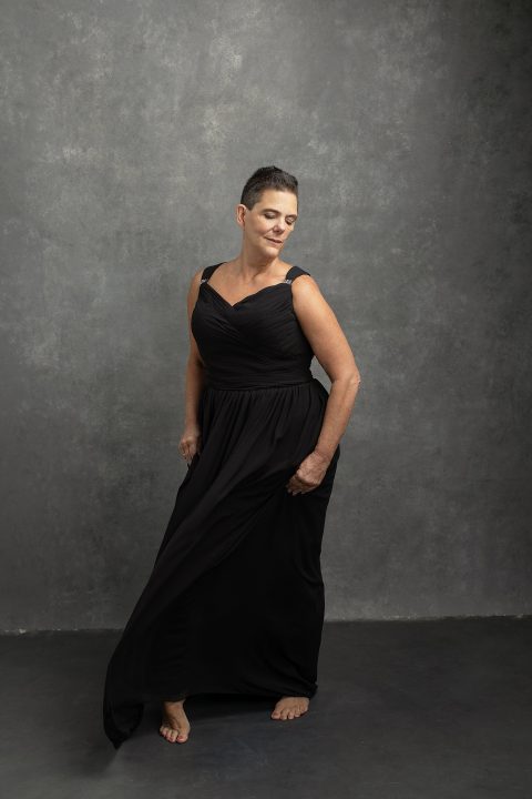 a portrait of a woman in her 50s, after cancer, wearing a long black dress, and looking down over her shoulder