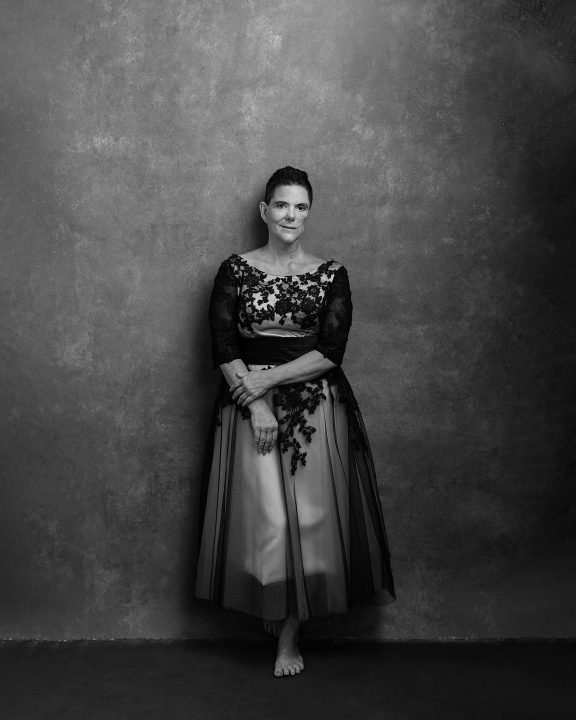 a black and white portrait of a woman in her 50s, after cancer, wearing a vintage lace dress, leaning against a wall with a gray hand-painted backdrop