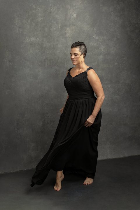 A portrait of a woman in her 50s, after cancer, wearing a long black dress and taking a step forward