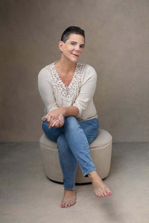 A casual portrait of a woman in her 50s with very short hair, wearing jeans and a cream-colored blouse, seated, against a beige backdrop