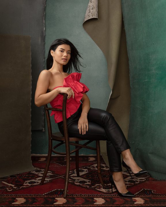 Portrait of a woman, seated, wearing a red ruffled top, with layered backdrops