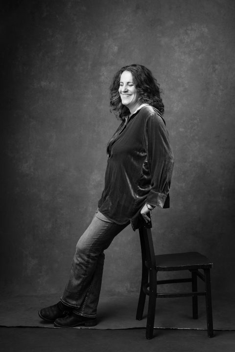 A black and white portrait of a woman laughing with her eyes closed, leaning on a chair